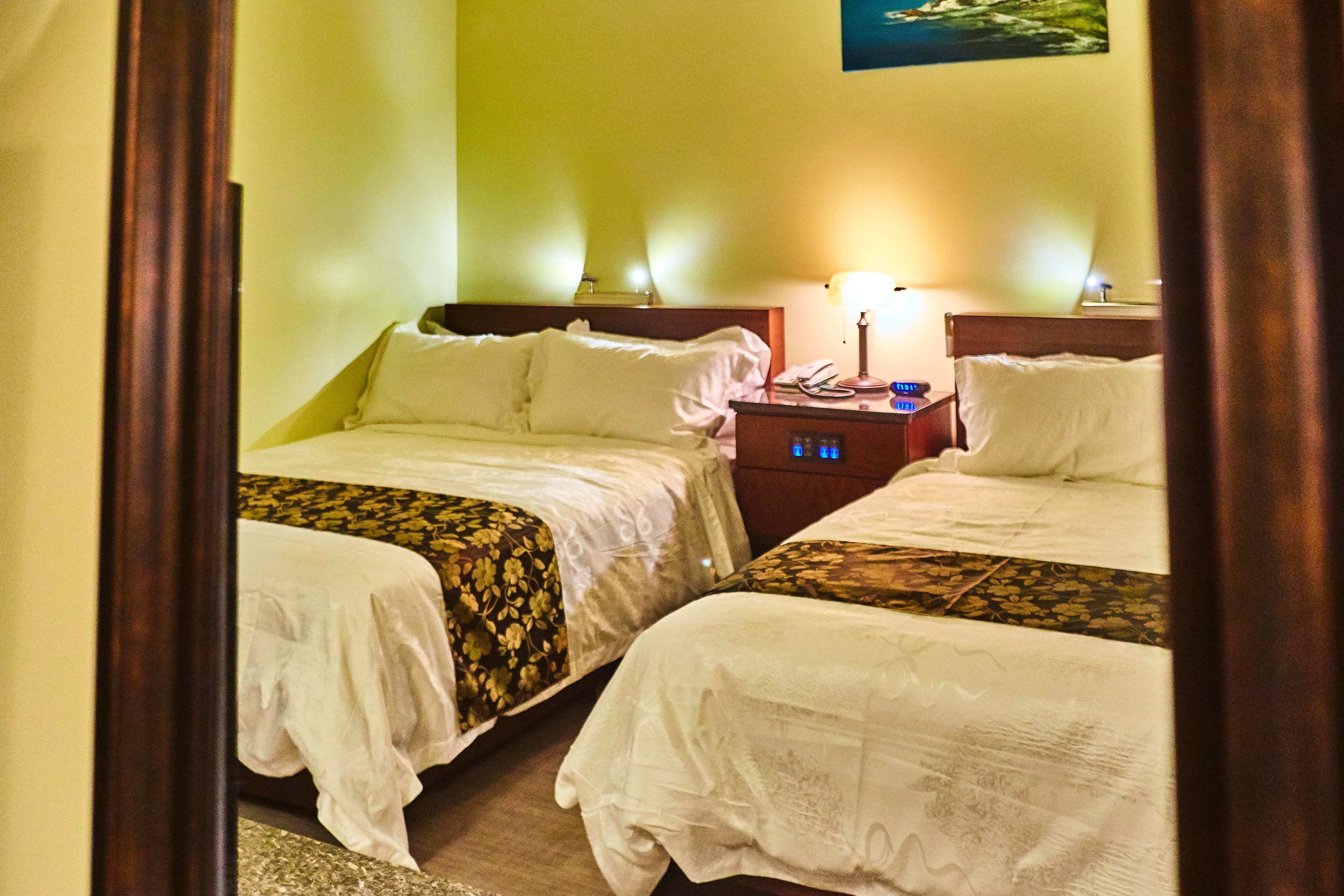 A hotel room with two beds, each with a floral-patterned bedspread and white pillows. There is a nightstand with a lamp and a phone between the beds.