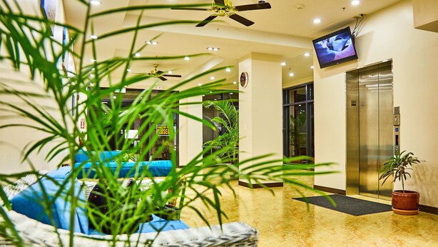 A lobby with a ceiling fan, a television mounted on the wall, an elevator, and a potted plant. - Serenti Hotel (Saipan)