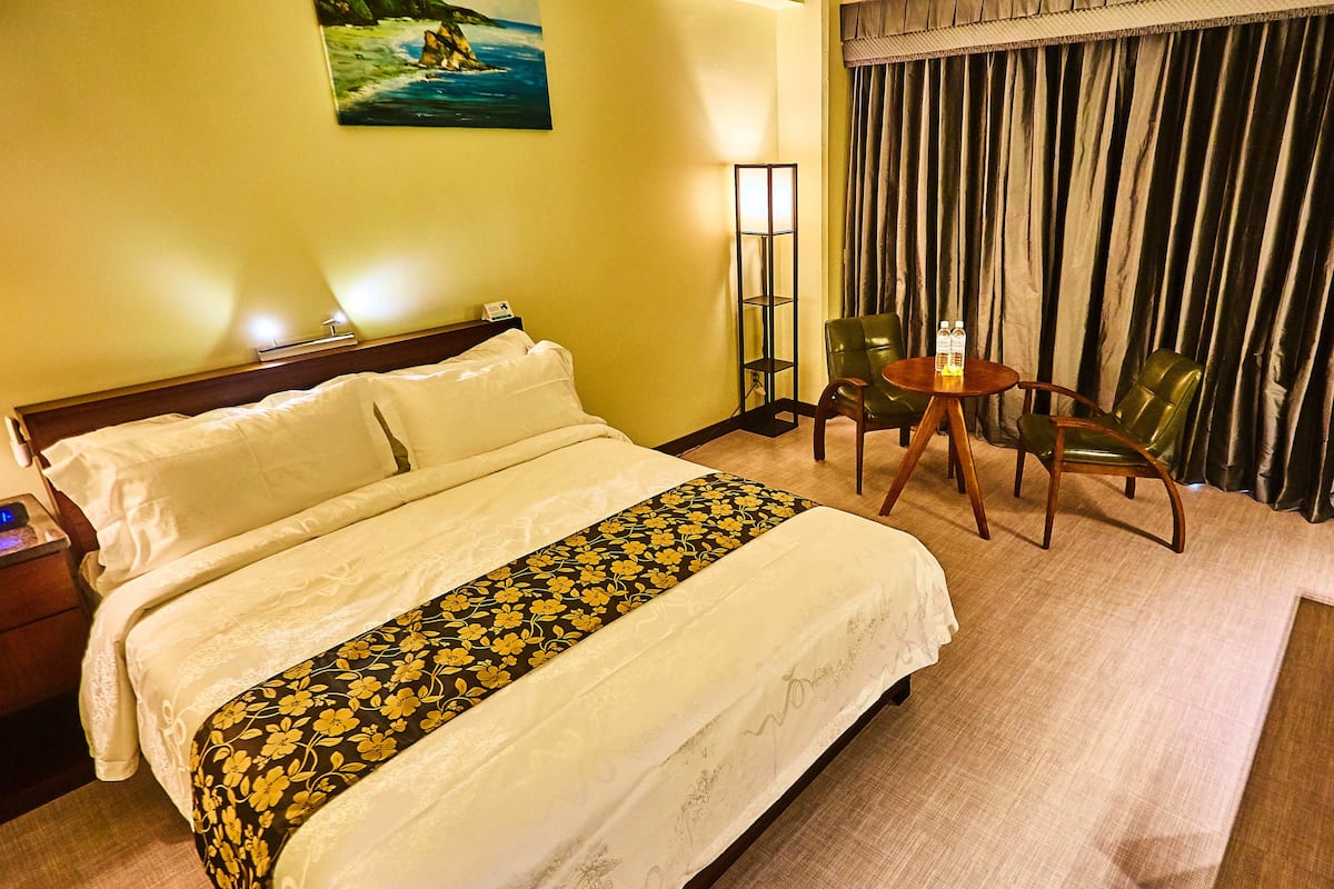 Une chambre d’hôtel avec un grand lit, une table de chevet avec une lampe, une chaise, une petite table avec une bouteille et un tableau au mur.