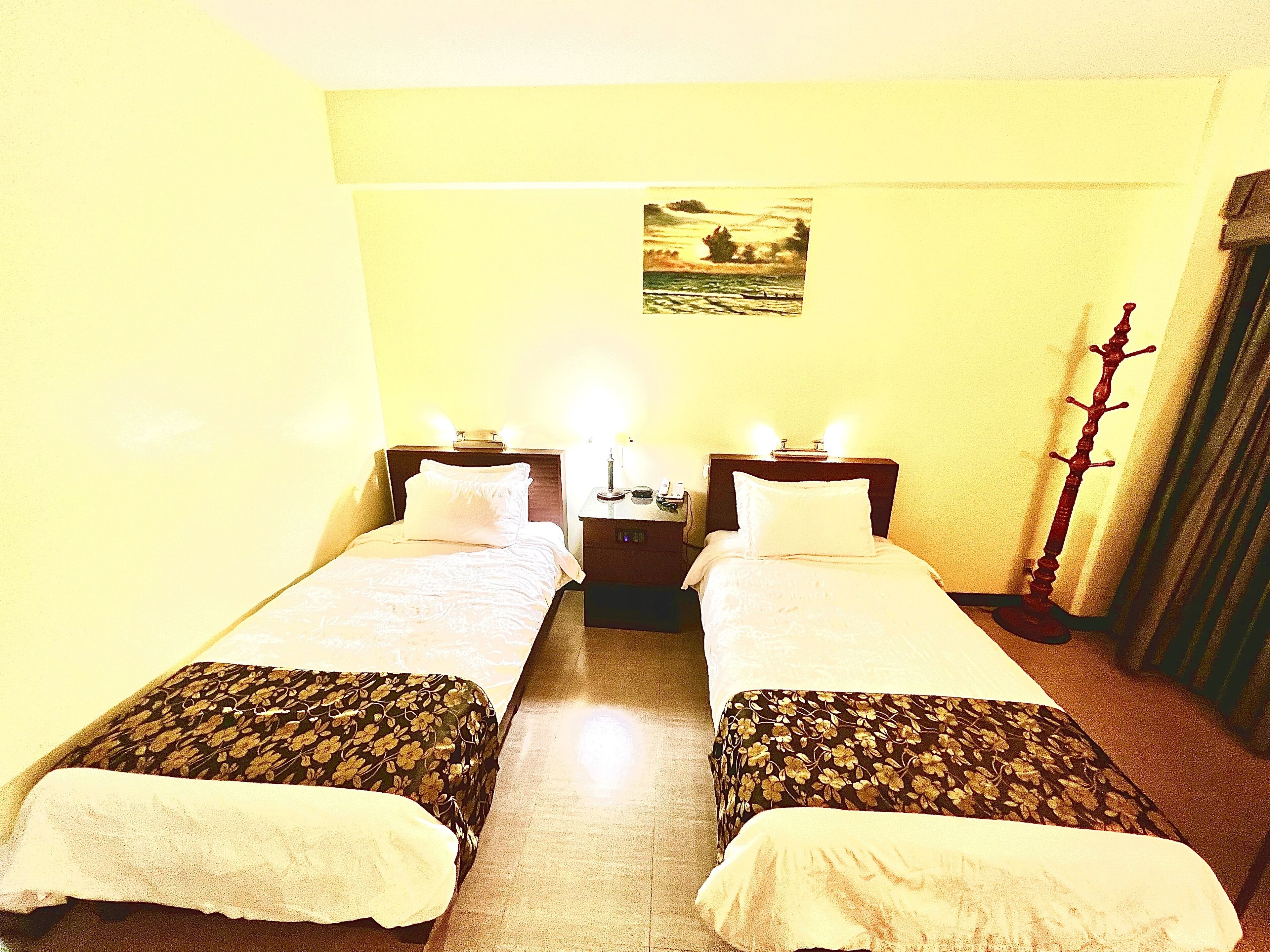 A double room with two beds, a nightstand with a lamp, a coat rack, and a painting on the wall.