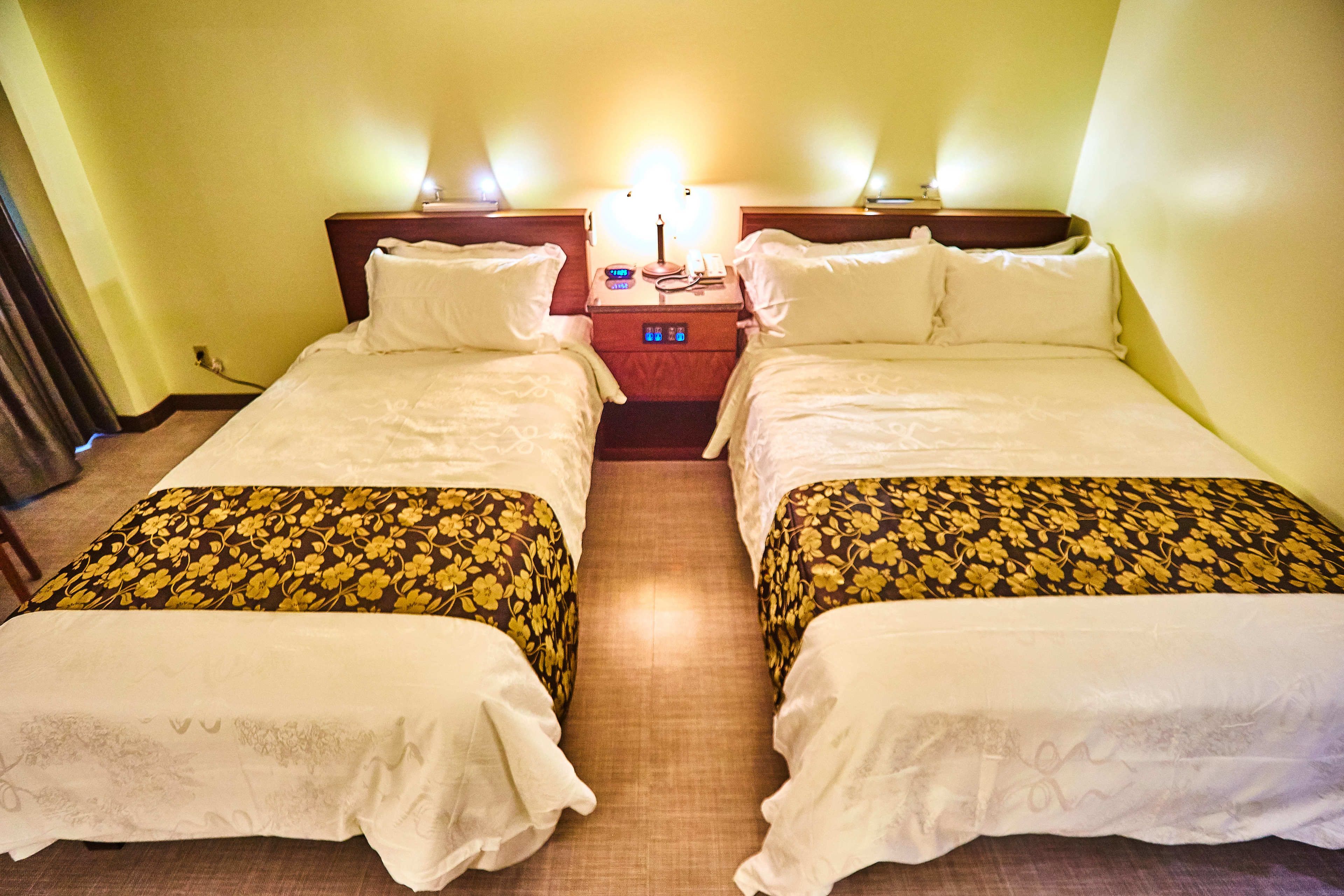 A hotel room with two beds, a nightstand with a lamp, and a wall-mounted clock.