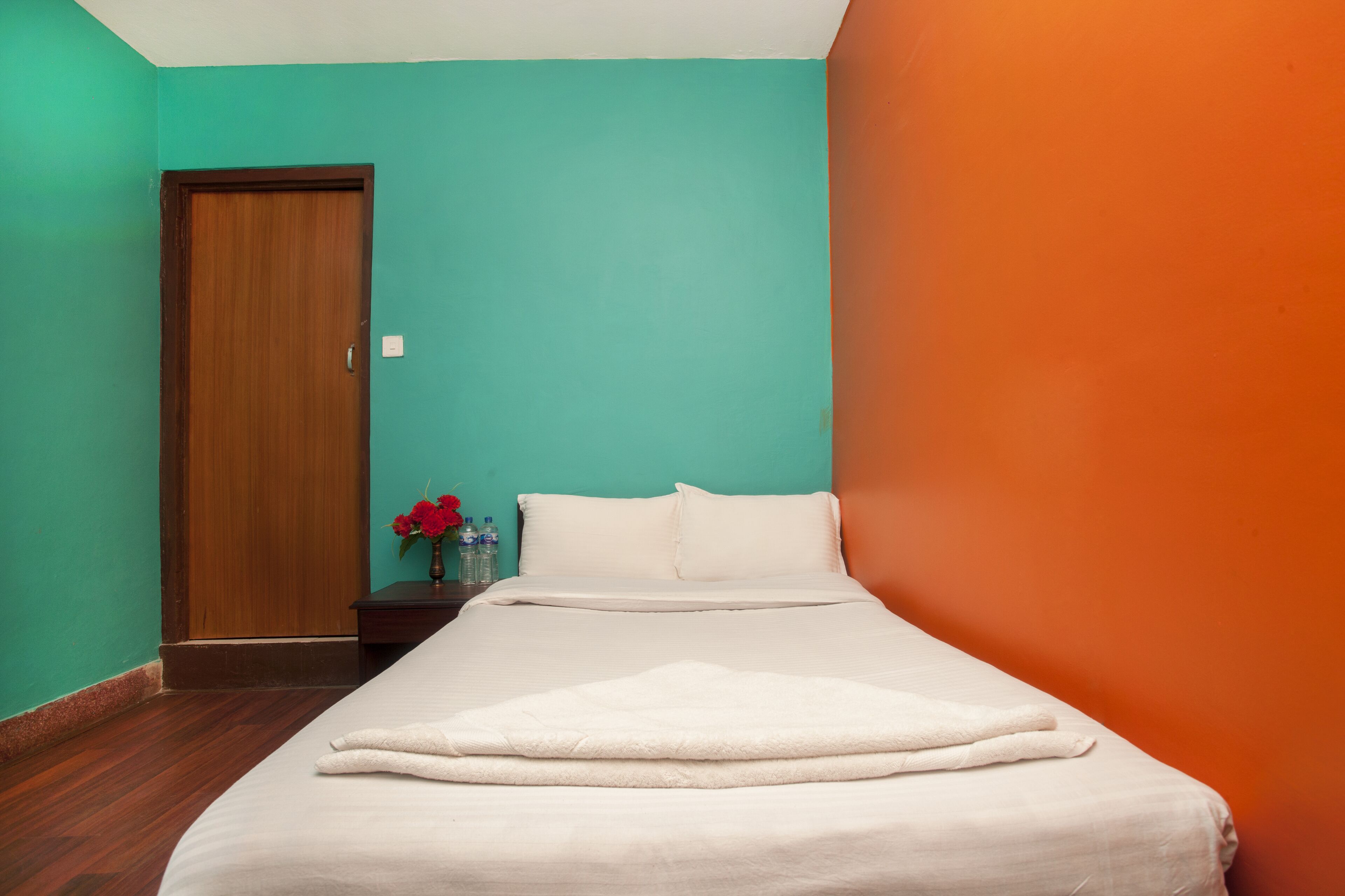Une chambre d’hôtel aux murs de couleur turquoise, avec une porte en bois, un lit recouvert de draps blancs et une petite table sur laquelle est posé un vase rempli de fleurs rouges.
