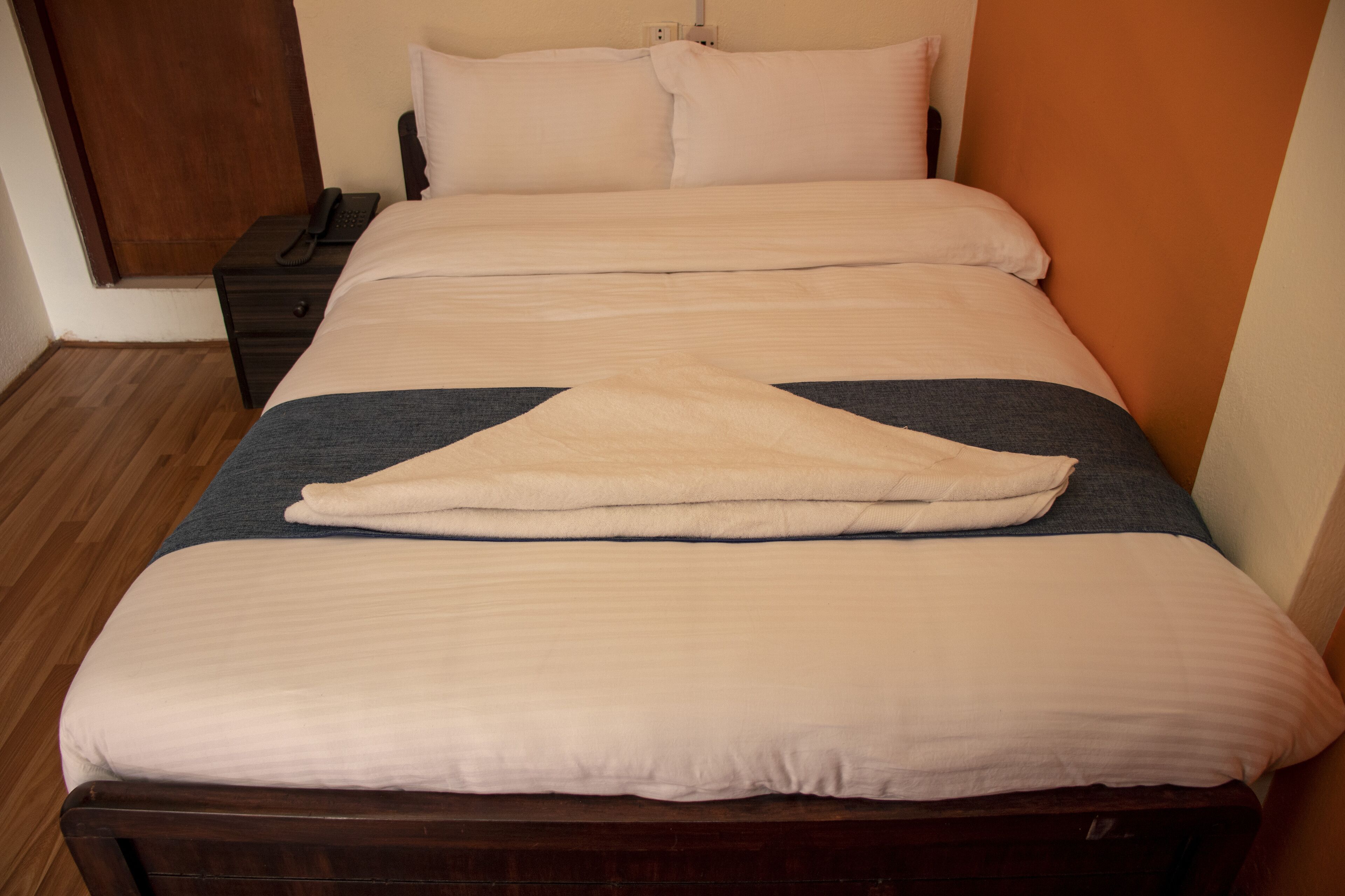 A neatly made bed with a dark headboard, a bedside table with a phone, and a wooden floor.