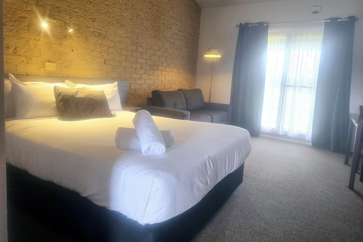 A hotel room with a brick wall, a bed with white linens, a grey sofa, and a large window with curtains.