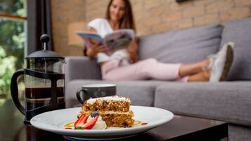 A plate of layered cake with fruit on a table, a coffee pot, and a person reading in the background.