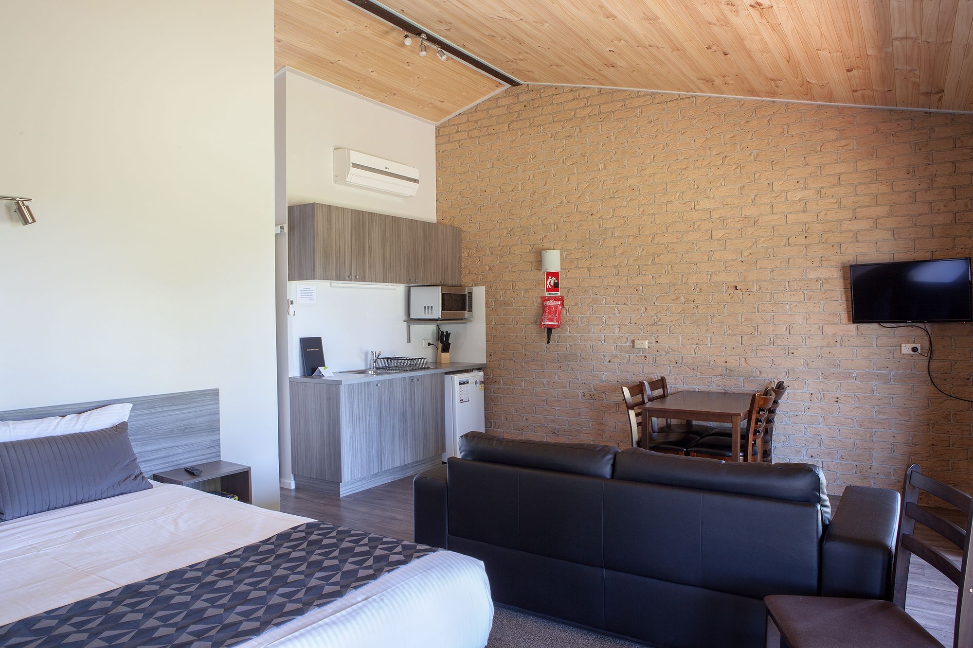 A compact hotel room with a bed, a sofa, a small kitchen area, and a television mounted on the brick wall.