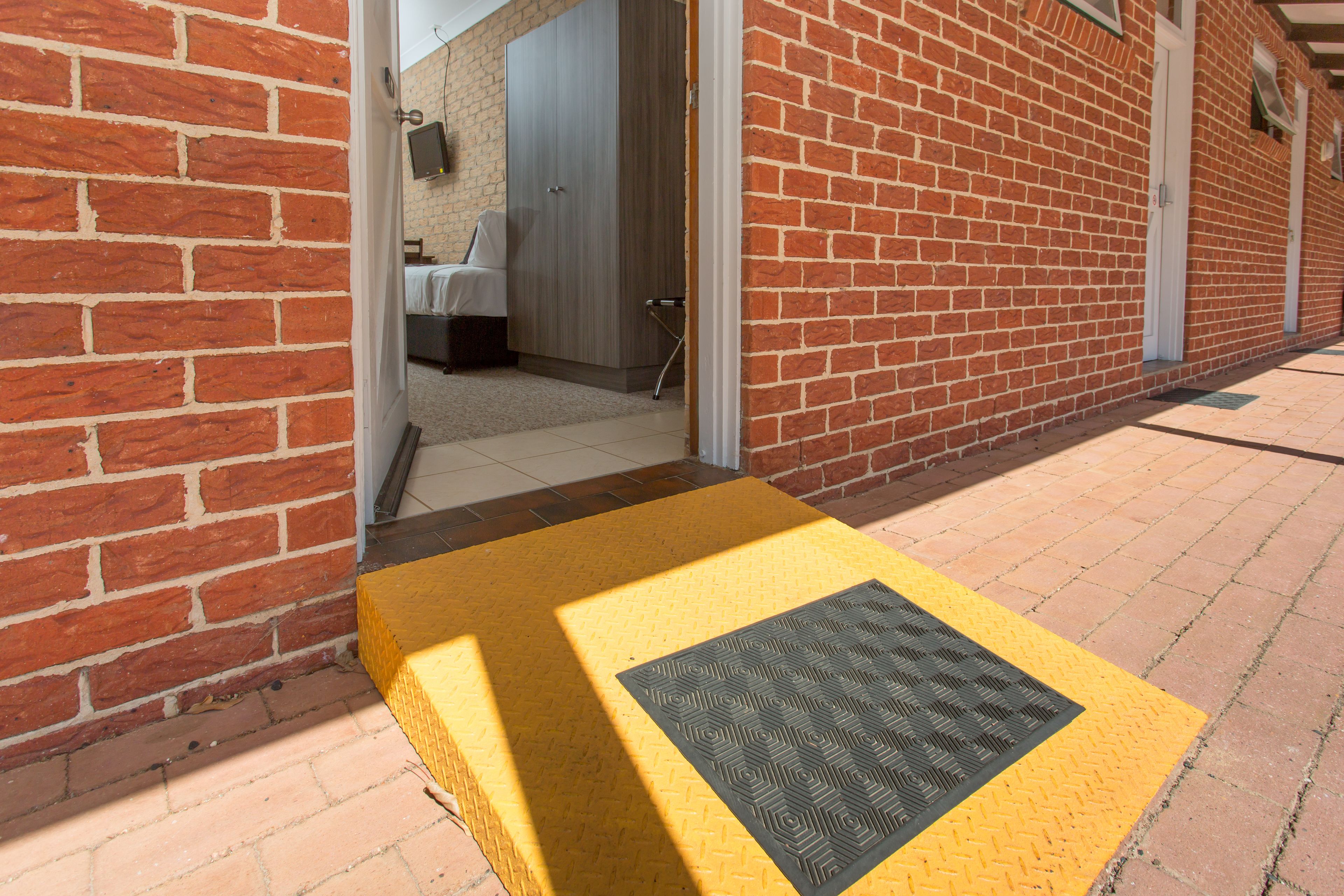 Entrada de hotel con un tapete podotáctil amarillo que conduce a una habitación con cama y armario.