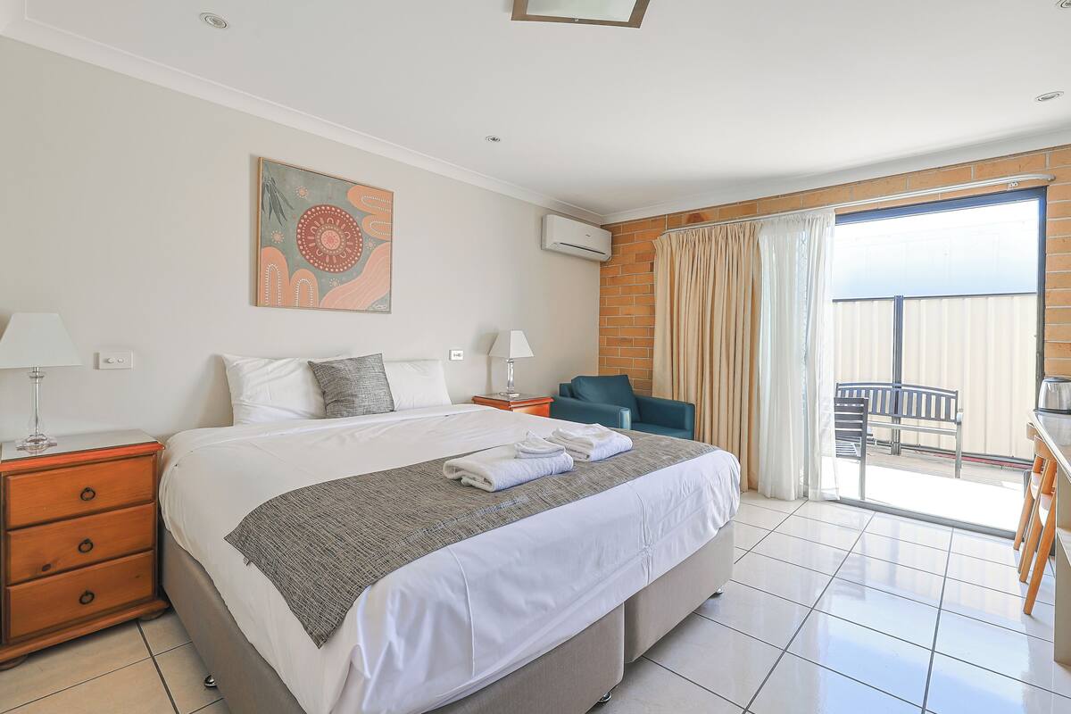 A hotel room with a large bed, a desk, a chair, a balcony with a view, and an air conditioning unit.
