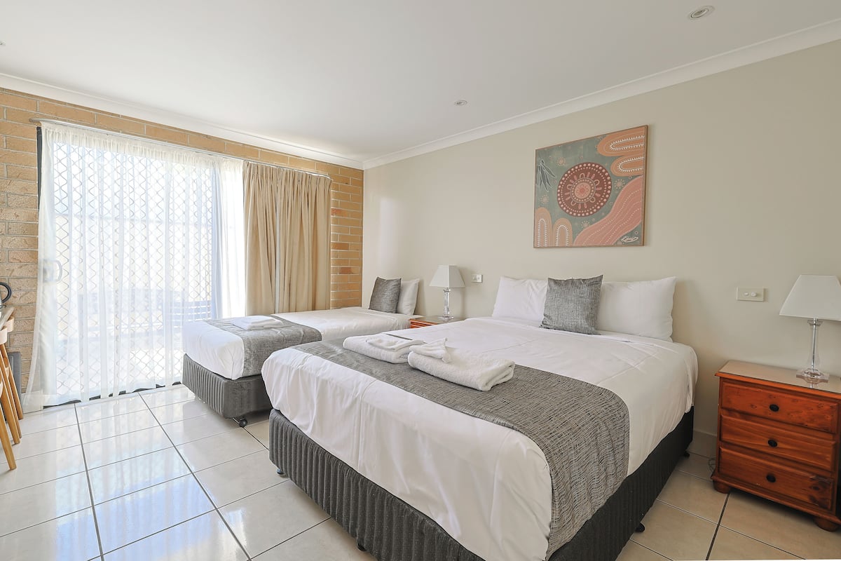A hotel room with two beds, a large window with curtains, a wooden nightstand, and a wall-mounted artwork.