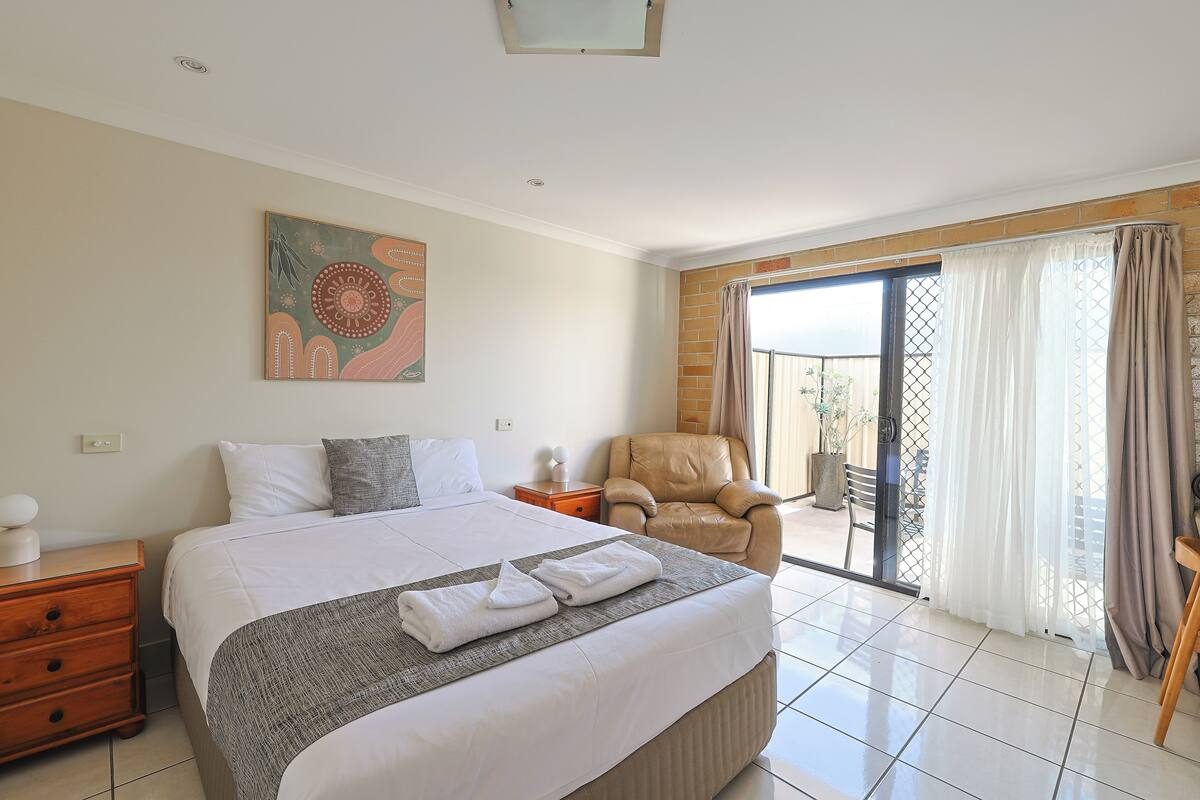 A hotel room with a large bed, a chair, a painting, and a view through a sliding door.