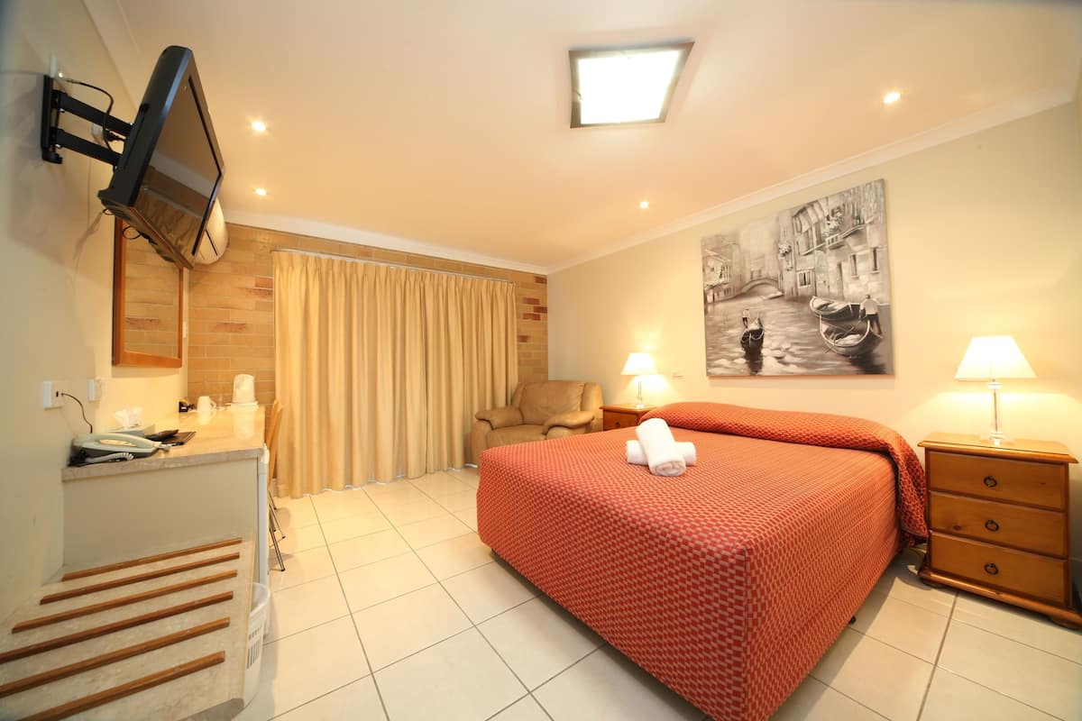 A hotel room with a bed, a desk, a chair, a television mounted on the wall, a painting, and a lamp.