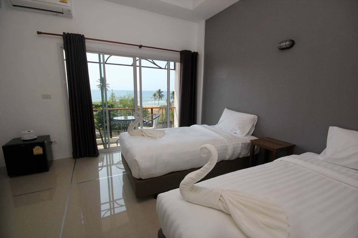 A hotel room with two beds, a small table, a nightstand, and a view of the beach and ocean through large windows.