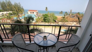 Un balcon avec deux fauteuils en osier et une table en verre, donnant sur une plage bordée de bâtiments et de palmiers.