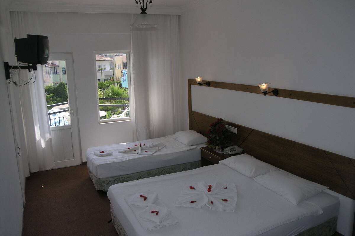 A hotel room with two beds, a TV, a window with curtains, and a view of greenery outside.