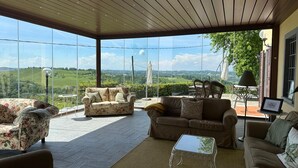 Un vaste espace de vie doté de grandes fenêtres offrant une vue panoramique sur des collines verdoyantes. Le mobilier comprend un canapé à motifs floraux, un canapé marron et une table basse en verre.