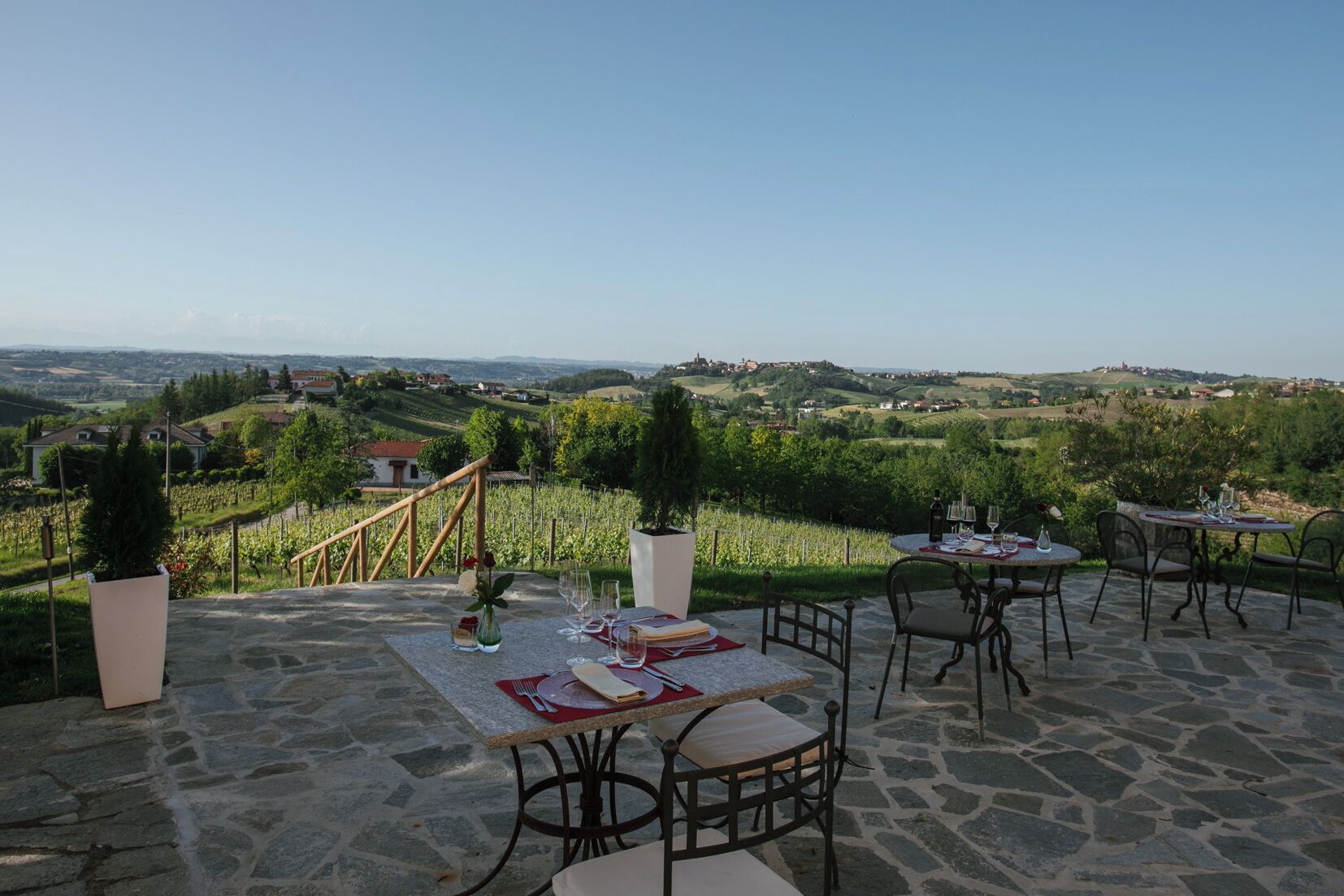 An outdoor dining area with stone flooring, tables set for two, and a scenic vineyard view.
