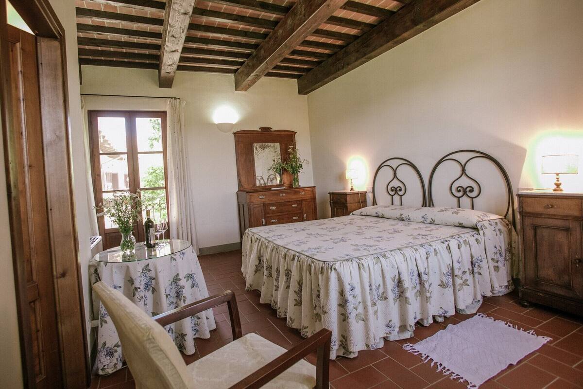 A bedroom with a bed, a wooden dresser, a nightstand, a chair, a table with a vase and wine glasses, and a window with curtains.