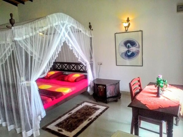 A room with a bed under a mosquito net, a table with a red tablecloth, a chair, a small cabinet, and a wall-mounted light fixture.