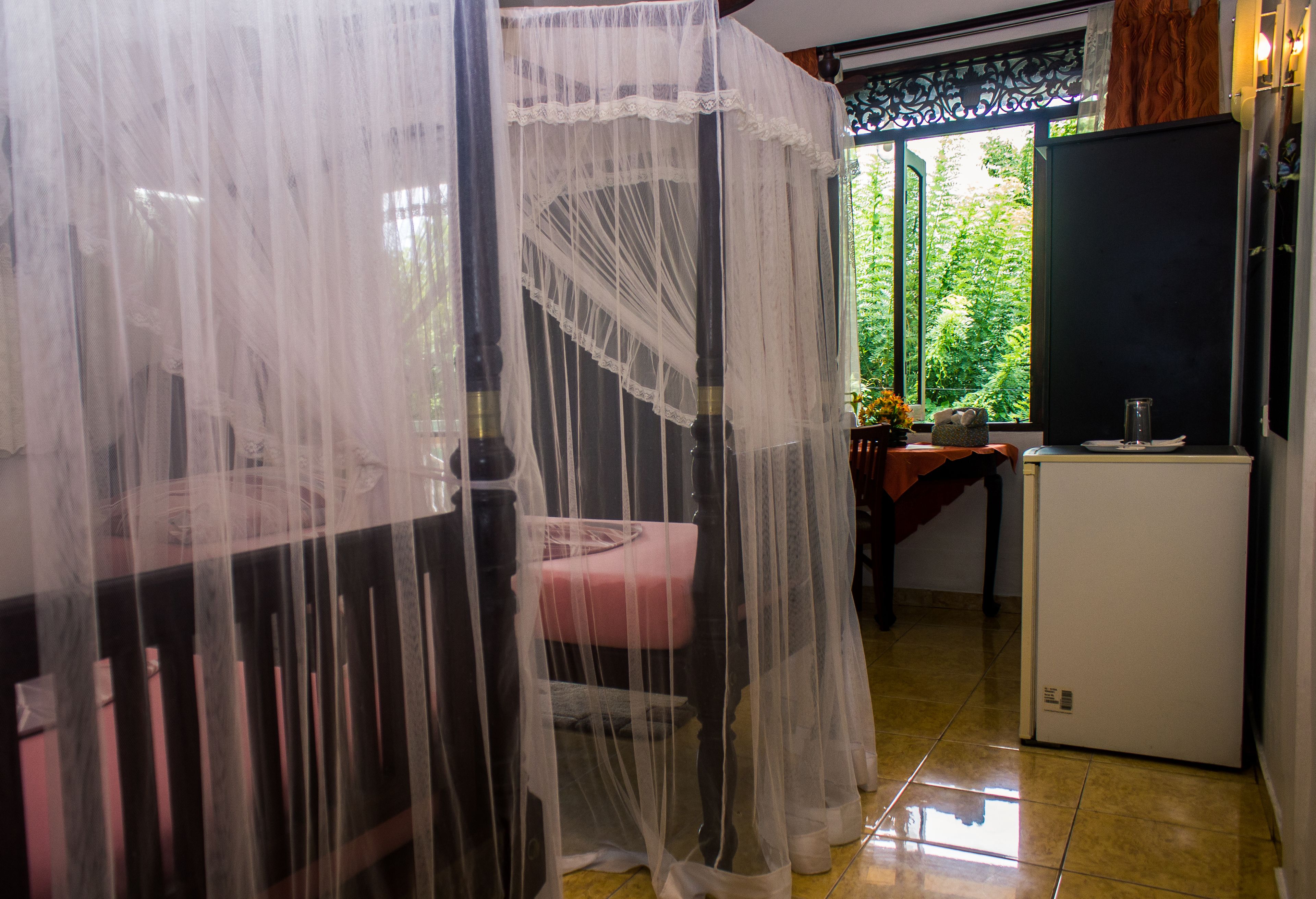 A room with a bed covered by a sheer canopy, a small table with a pink cushion, a refrigerator, and a window with a view of greenery.
