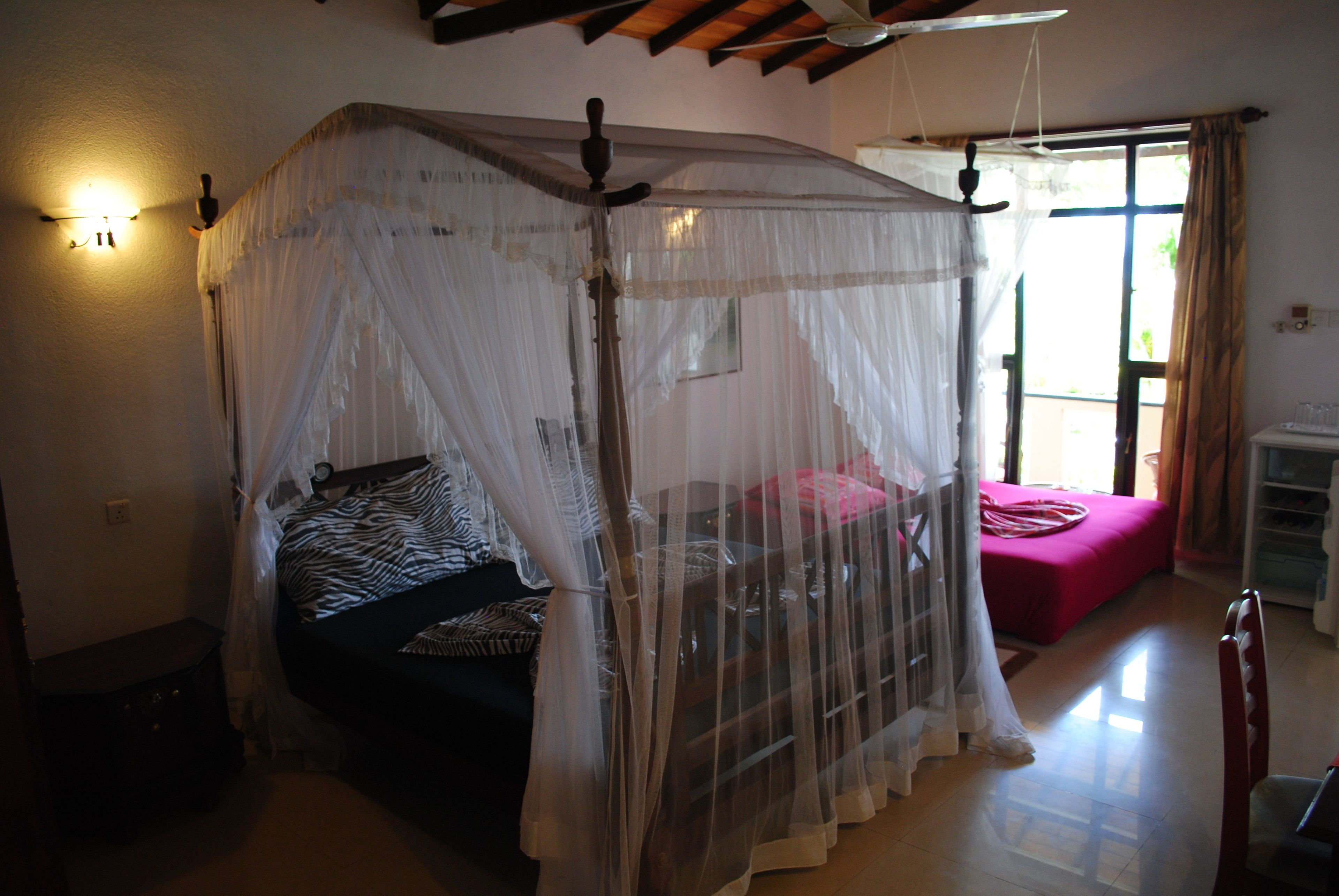 A bedroom with a canopy bed, a small table, a chair, a window with curtains, and a view of the outside.