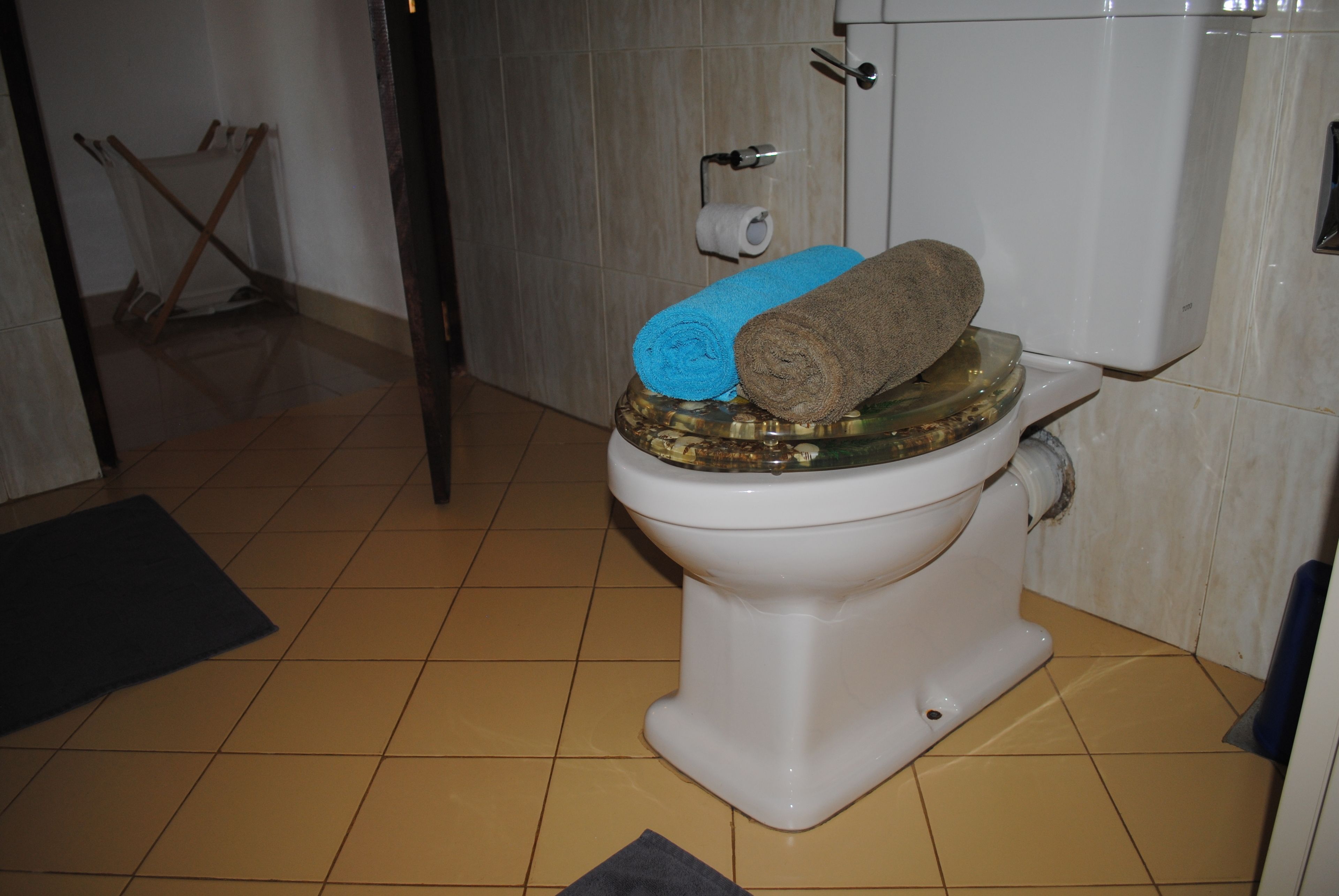 A toilet with a blue and brown towel on top, a roll of toilet paper on the wall, and a broom leaning against the wall.