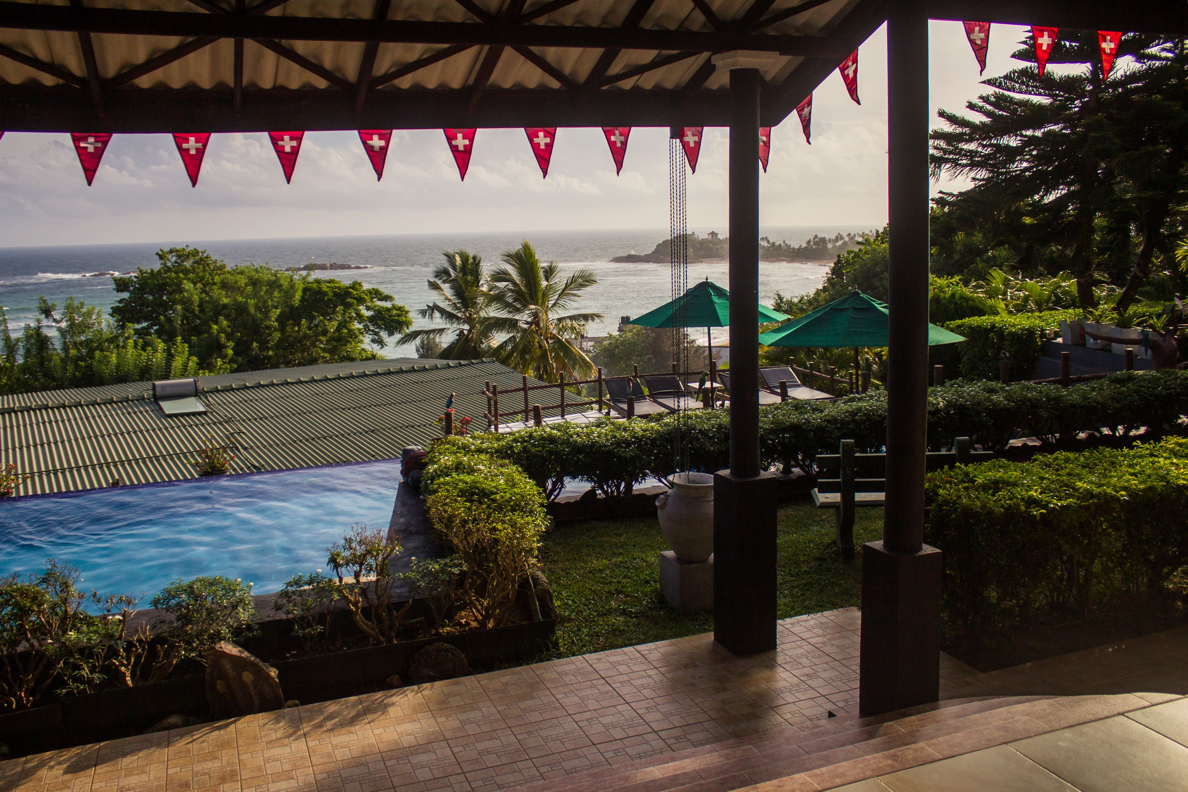 A pool area with a view of the ocean, surrounded by greenery and umbrellas.