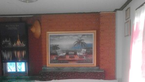 A hotel room with a bed, a painting of a beach scene, a window with a view of palm trees, and a small table with a lamp. - View Point Lodge (Mae Hong Son)