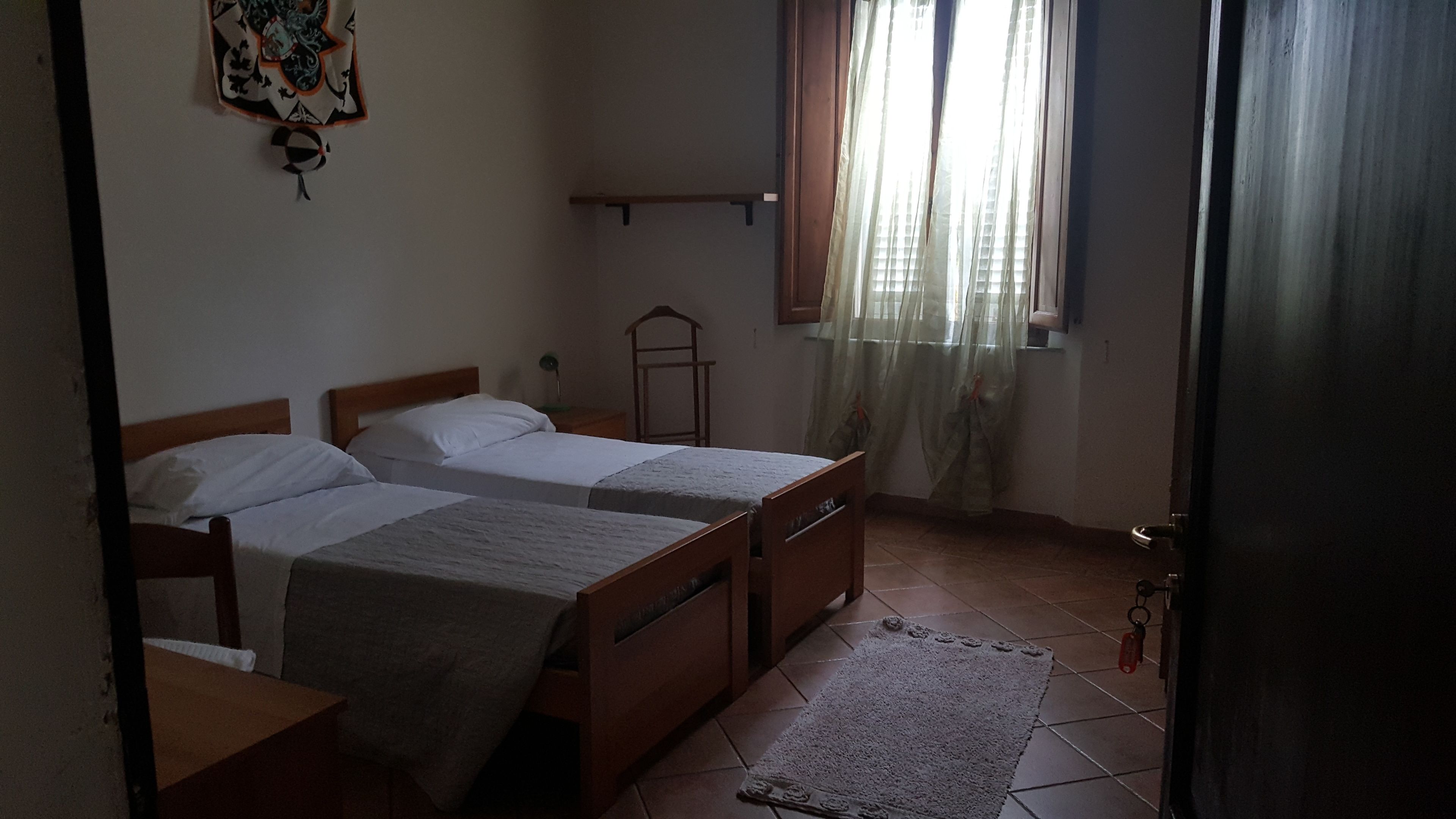 A bedroom with two single beds, a small table, a chair, a window with curtains, and a radiator.