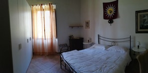 Quarto com cama, uma cadeira, mesa pequena, mesa de cabeceira, cortina, quadro na parede e uma bandeira.