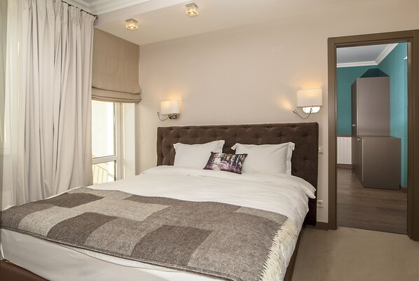 Habitación de hotel con una cama grande, dos lámparas de noche, una ventana con cortinas y vistas a un pasillo y un refrigerador.