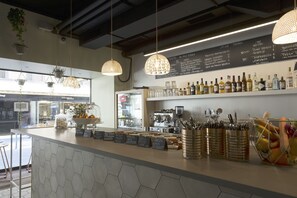Interior de una cafetería con un mostrador que exhibe diversos alimentos y bebidas, luces colgantes y una pizarra con el menú.