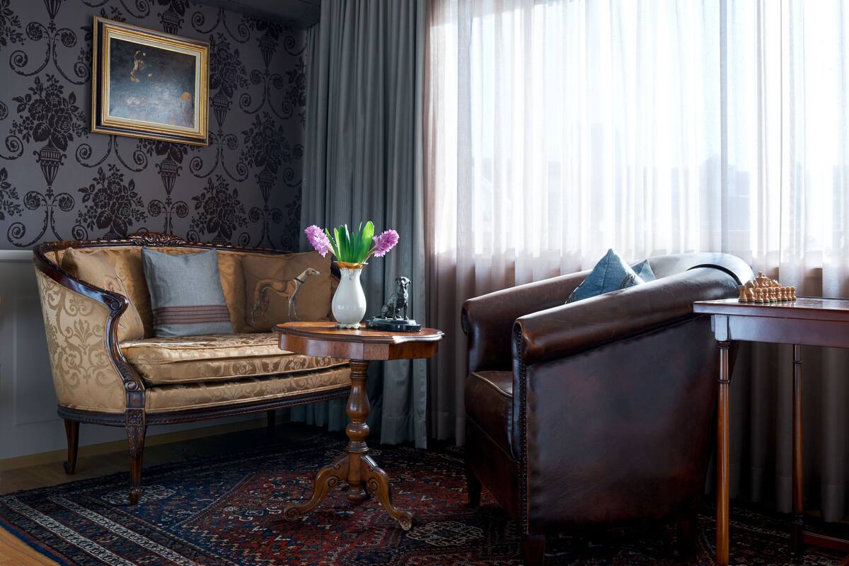 Ein Zimmer mit einem Vintage-Sofa, einem kleinen runden Tisch mit Blumen in einer Vase und einem Ledersessel.