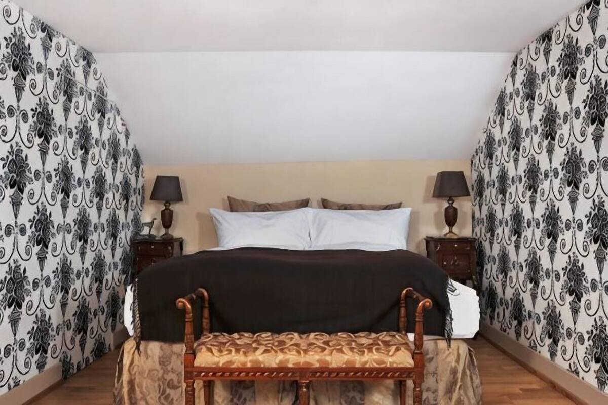 Ein Bett mit Kopfteil, zwei Nachttische mit Lampen, eine Bank und Wandtapete mit Muster.
