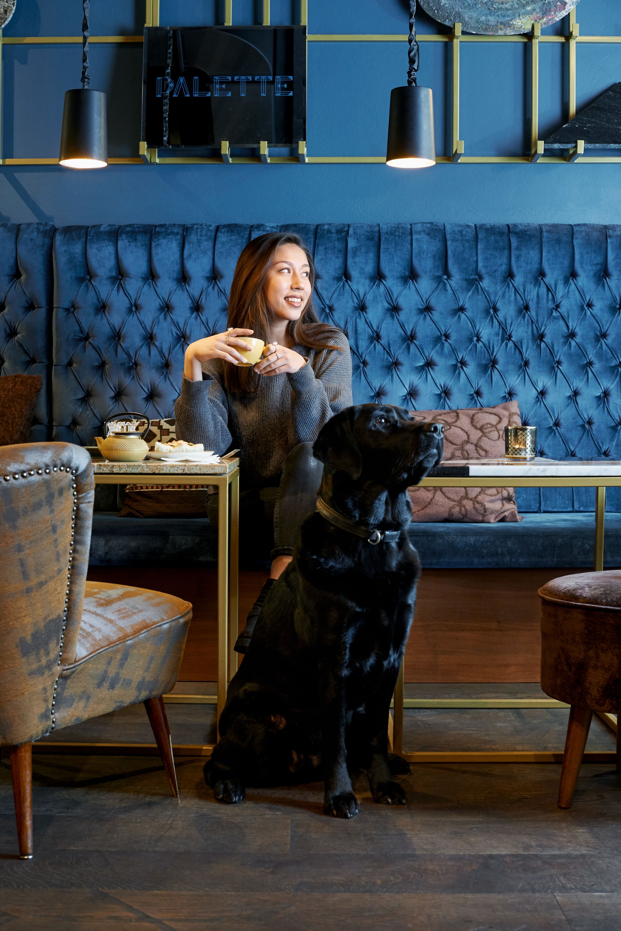 Una mujer sentada a una mesa con un perro, un sofá tapizado en azul y un letrero de neón en la pared que dice "PALETTE".