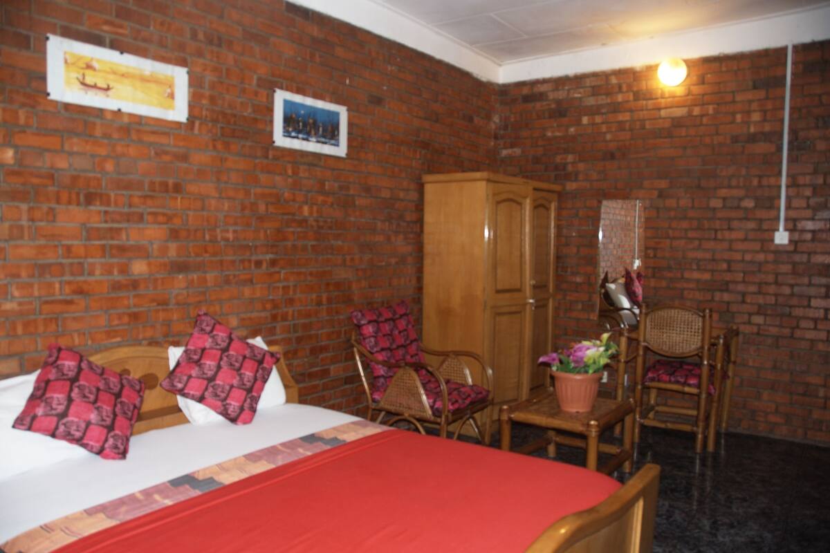 A room with a brick wall, a wooden wardrobe, two wicker chairs, a small table with a potted plant, and a bed with red bedding and patterned pillows.