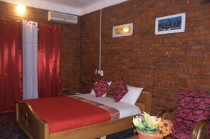 A room with a brick wall, a bed with red bedding, a wooden headboard, a potted plant, and a picture on the wall. - Joy Family Lodge (Amasaman)