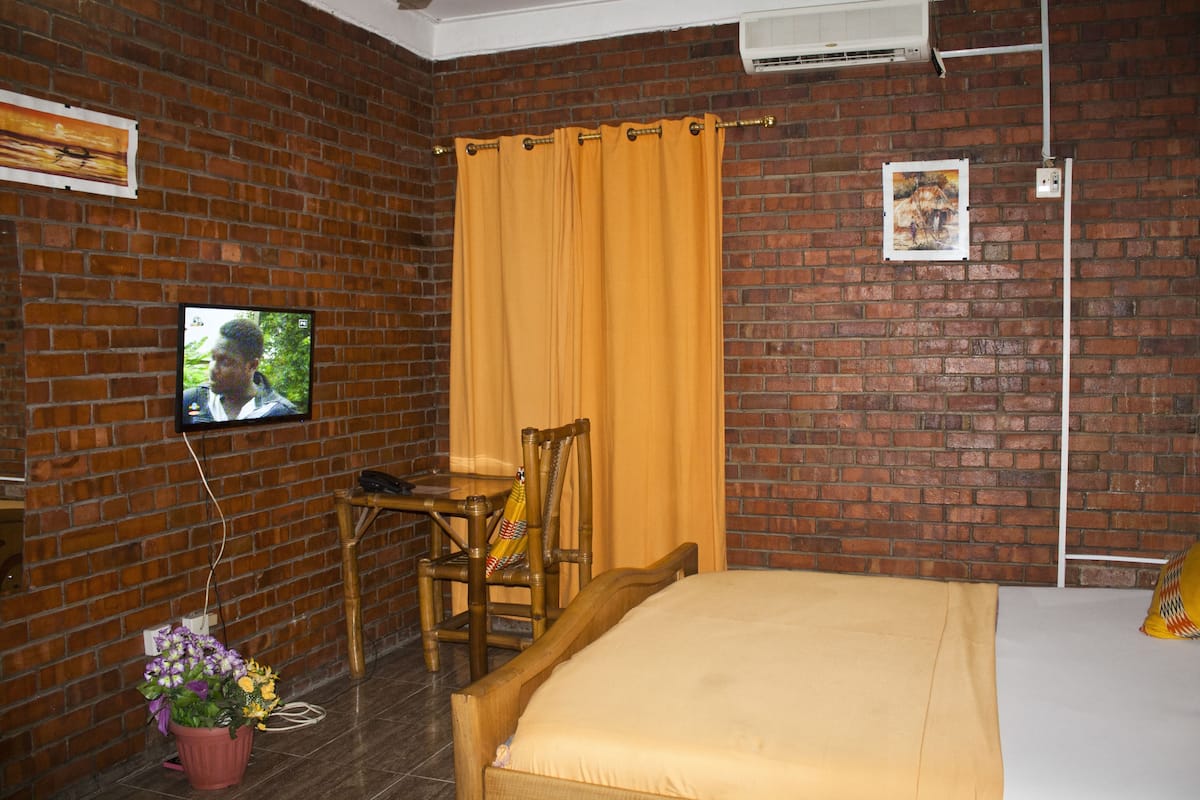 A room with a brick wall, a bed with a beige bedspread, a wooden chair, a small table, a television mounted on the wall, and a potted plant.