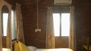 A room with a brick wall, a bed with white bedding and yellow pillows, a wooden chair, and two windows with curtains. - Joy Family Lodge (Amasaman)