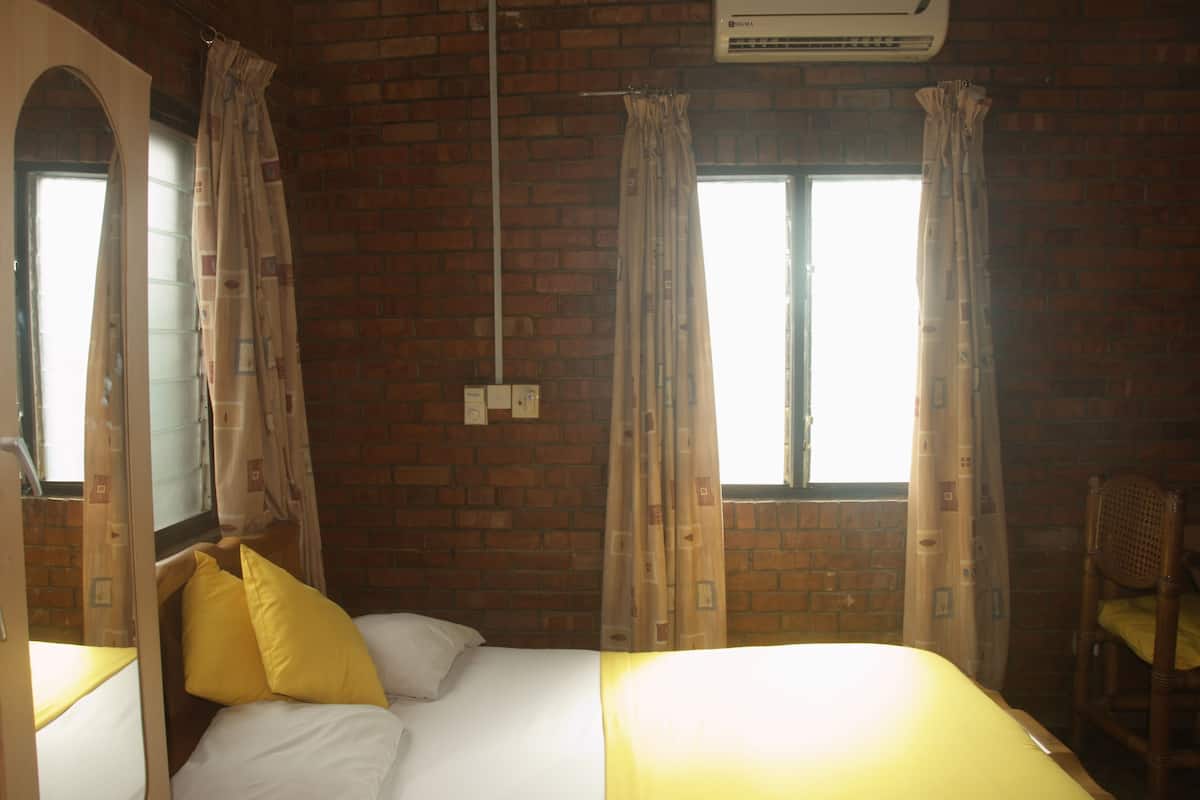 A room with a brick wall, a bed with white bedding and yellow pillows, a wooden chair, and two windows with curtains.