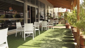 An outdoor patio with white wicker chairs and tables, surrounded by potted plants and a wooden pergola. - Ugur Hotel (Mersin)