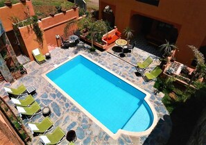 Una piscina rettangolare con bordo in pietra, circondata da poltrone a sdraio e tavolini, e una piccola zona salotto con divano e tavolo.