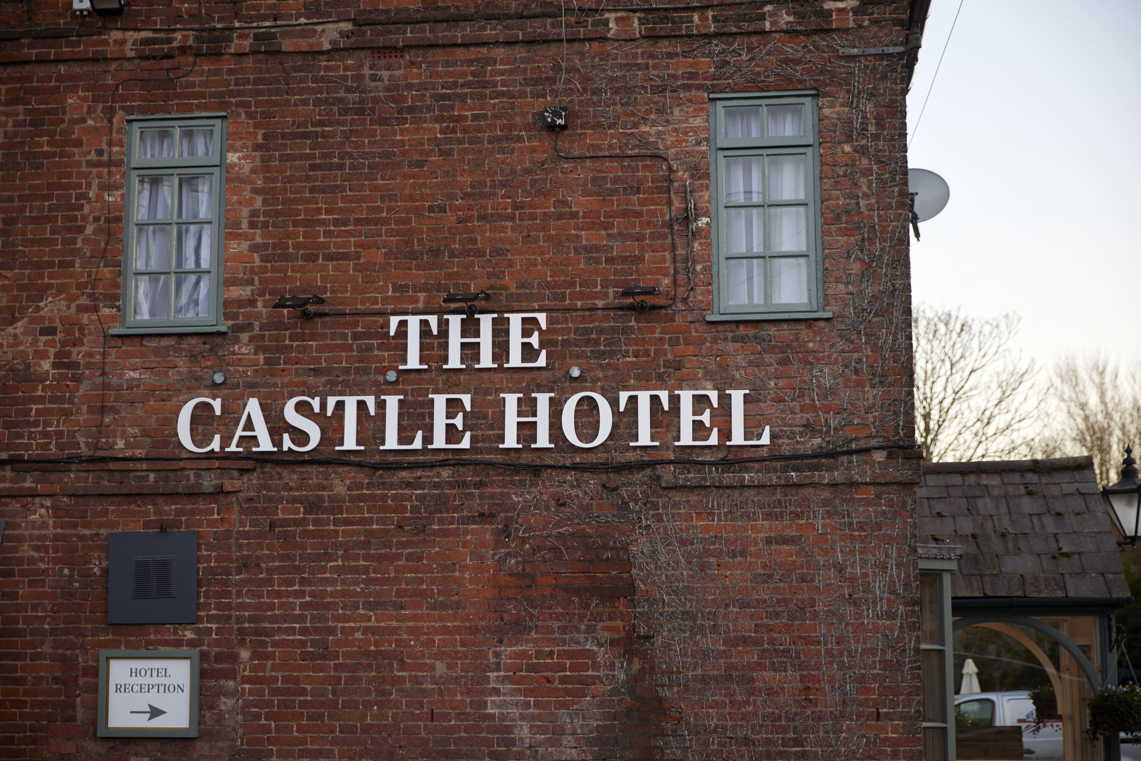 A brick building with a sign that reads "The Castle Hotel." There is a sign indicating the hotel reception and a satellite dish on the roof.