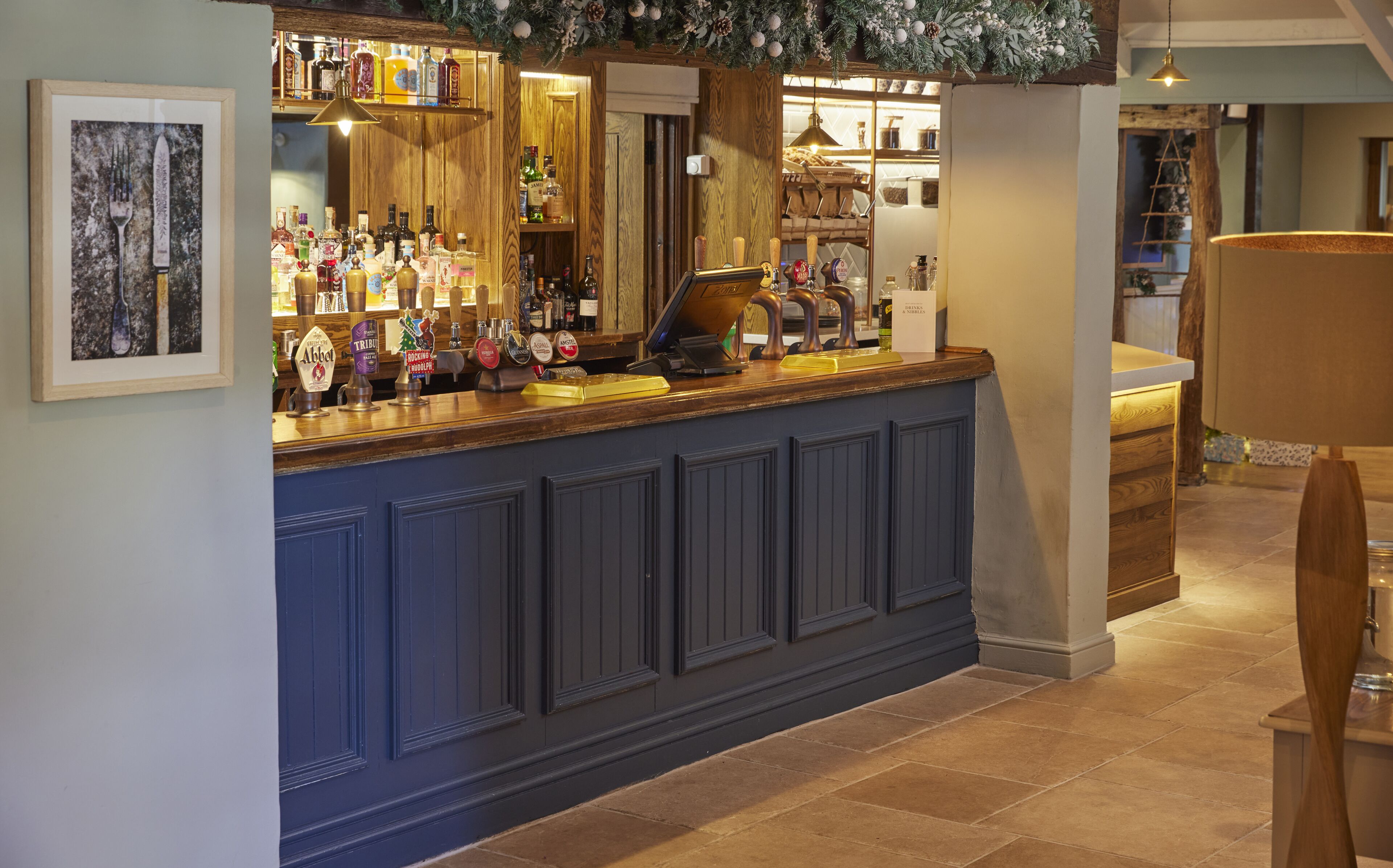 A bar area with a wooden counter, various bottles, and a framed artwork on the wall.