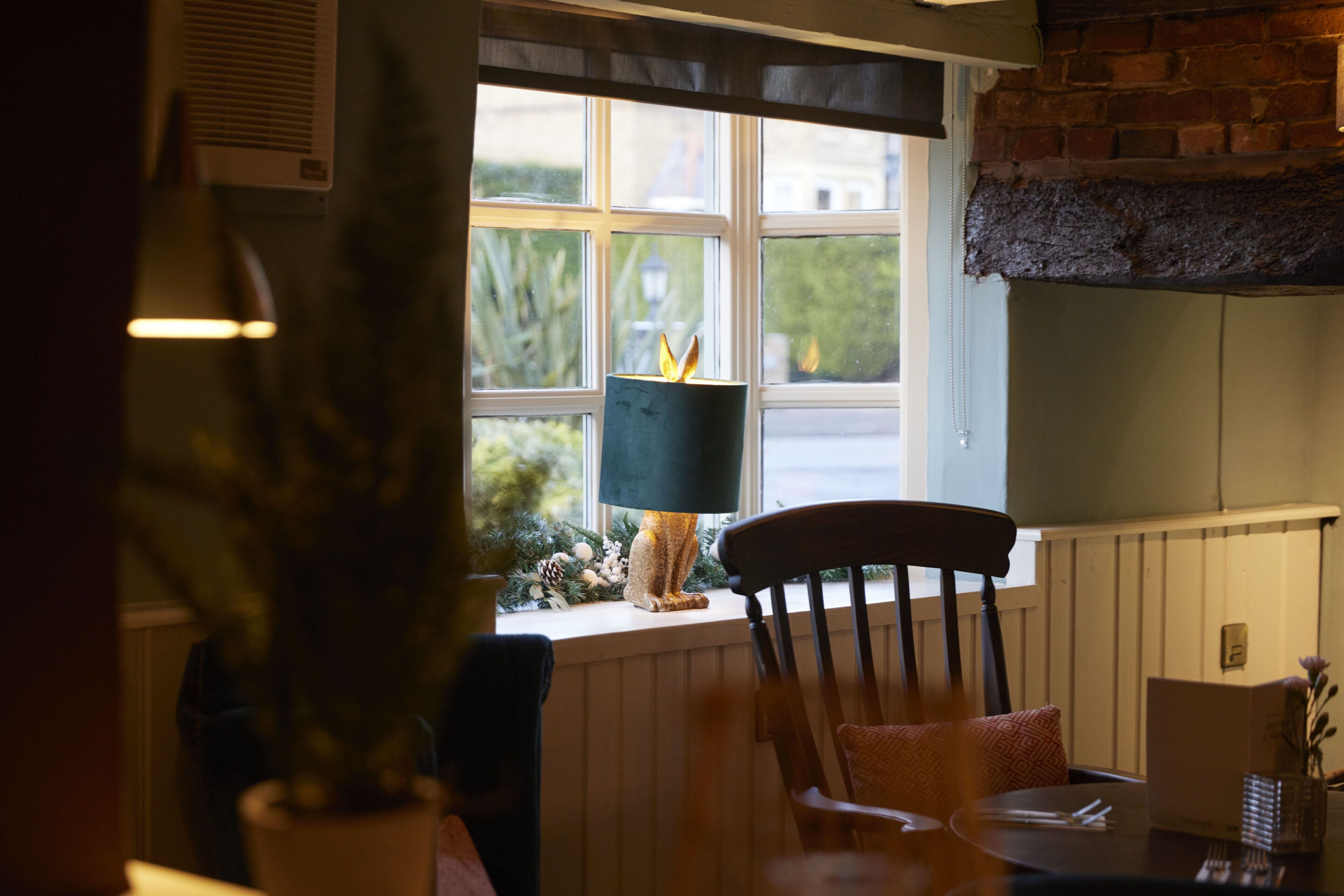 Un espace repas comprenant une table, des chaises, une fenêtre donnant sur un paysage verdoyant, une lampe décorative et un mur en briques.