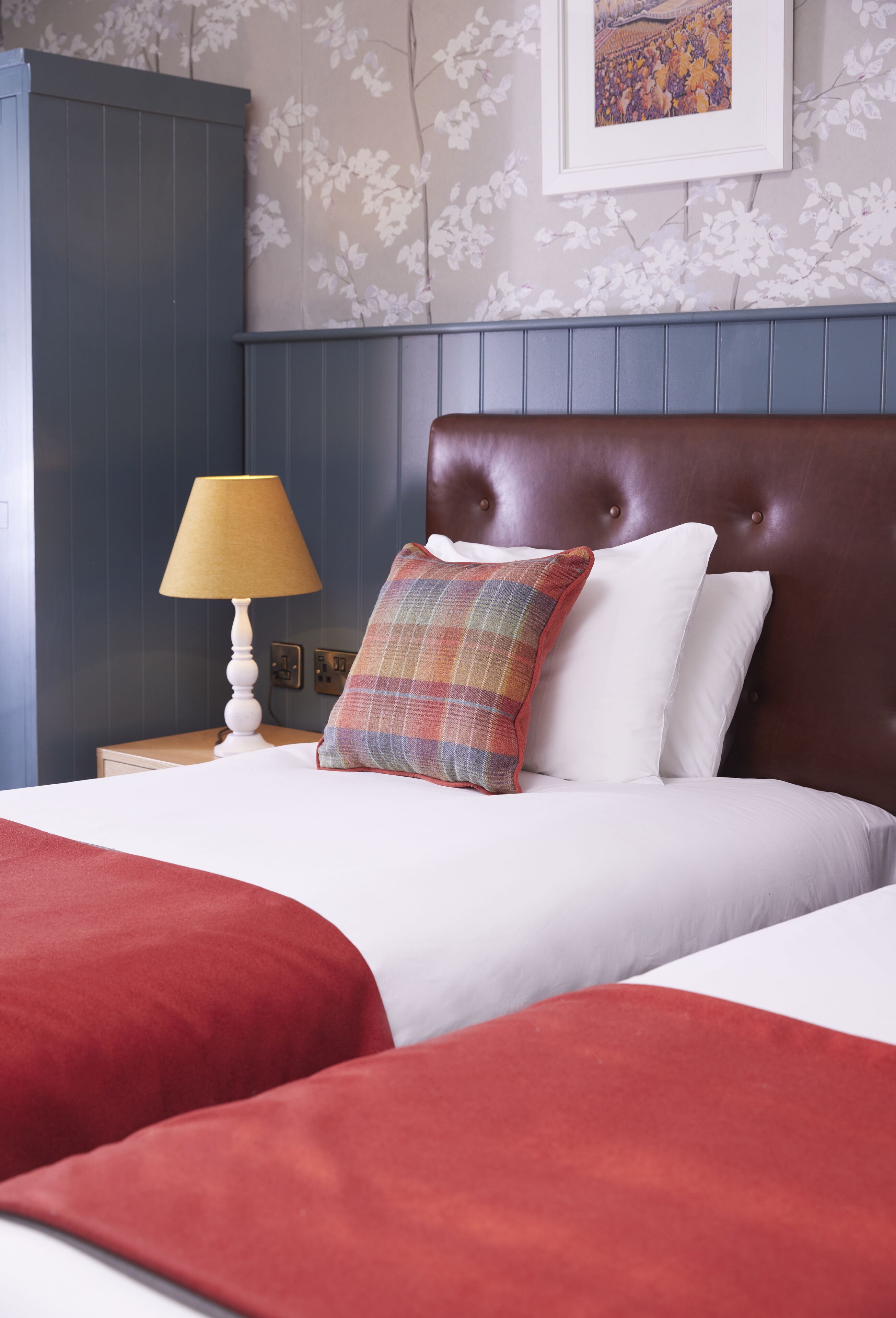 A neatly made bed with white linens and a plaid throw pillow, a wooden nightstand with a lamp, and a framed picture on the wall.