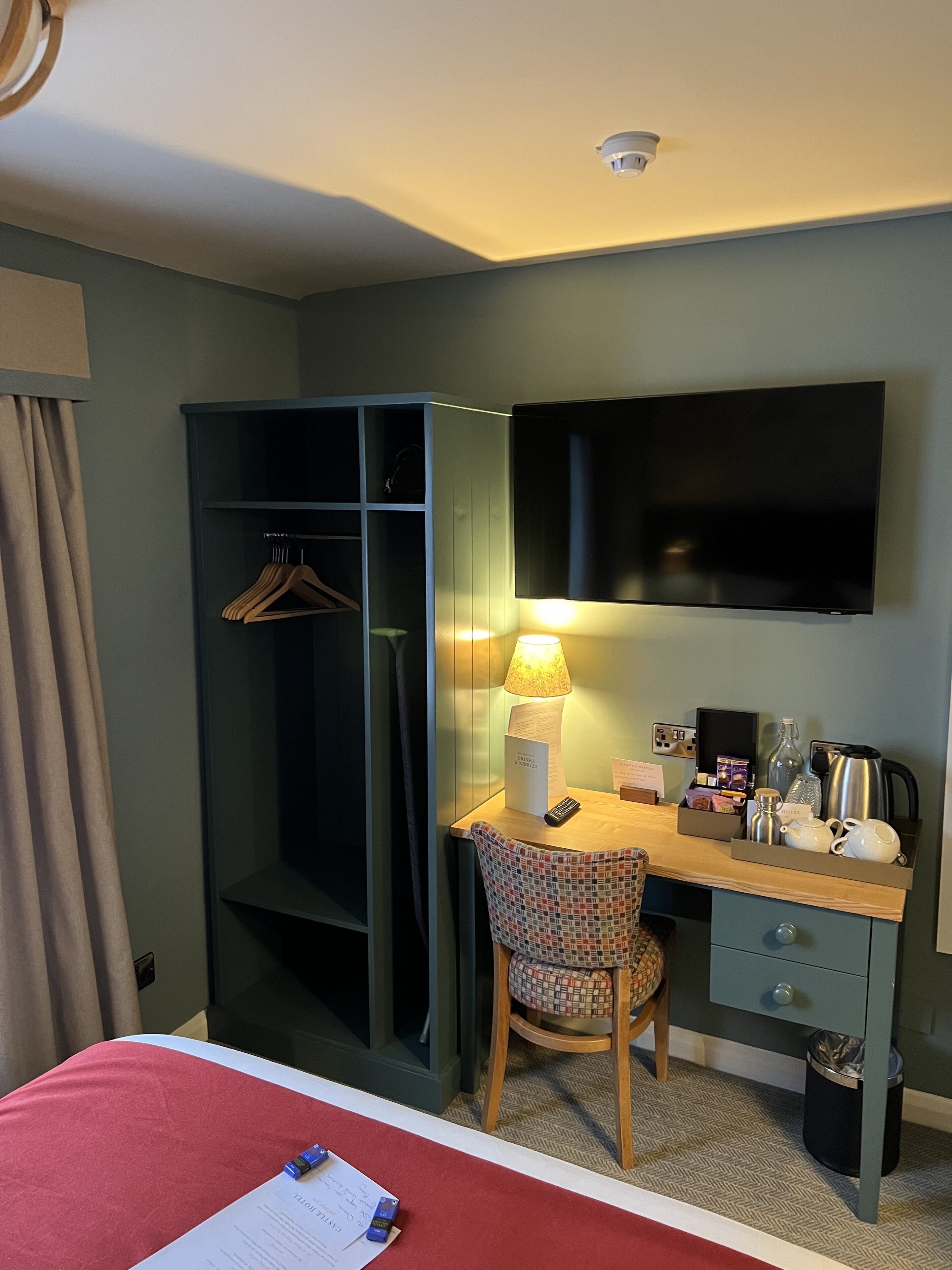 A hotel room with a bed, a desk with a lamp, a chair, a wardrobe, and a TV on the wall.