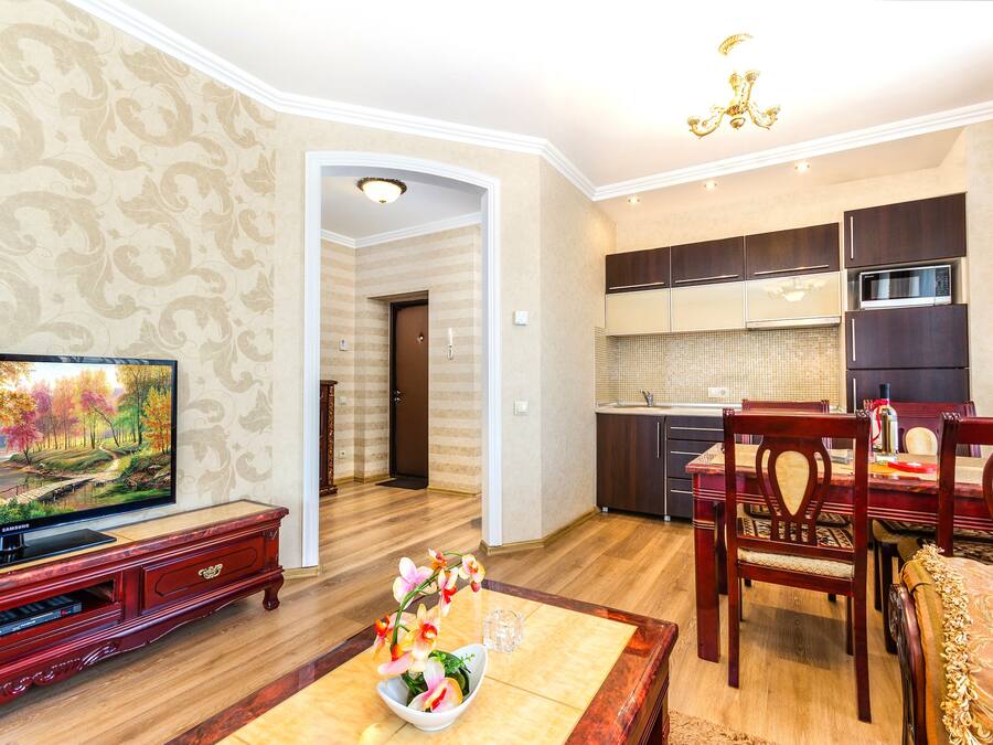 A living room with a flat-screen TV, wooden entertainment unit, dining table with chairs, and a kitchen area with modern appliances.