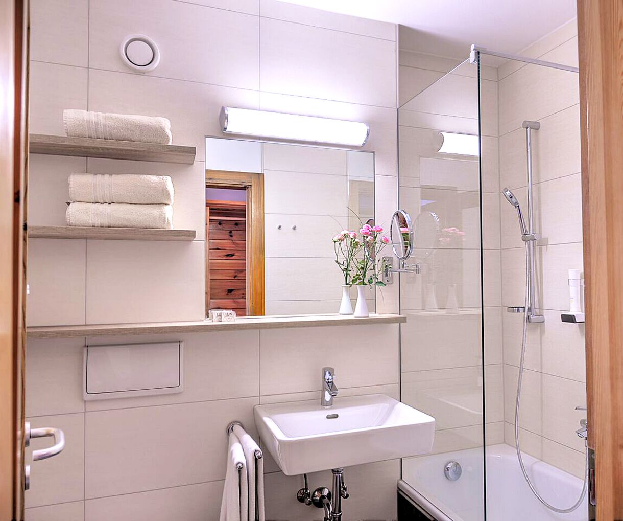 A modern bathroom with a glass-enclosed shower, a white sink, and a mirror above it. There are towels on a shelf and a vase with flowers on the countertop.