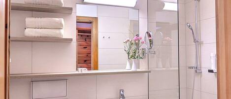 A modern bathroom with a glass-enclosed shower, a white sink, and a mirror above it. There are towels on a shelf and a vase with flowers on the countertop.