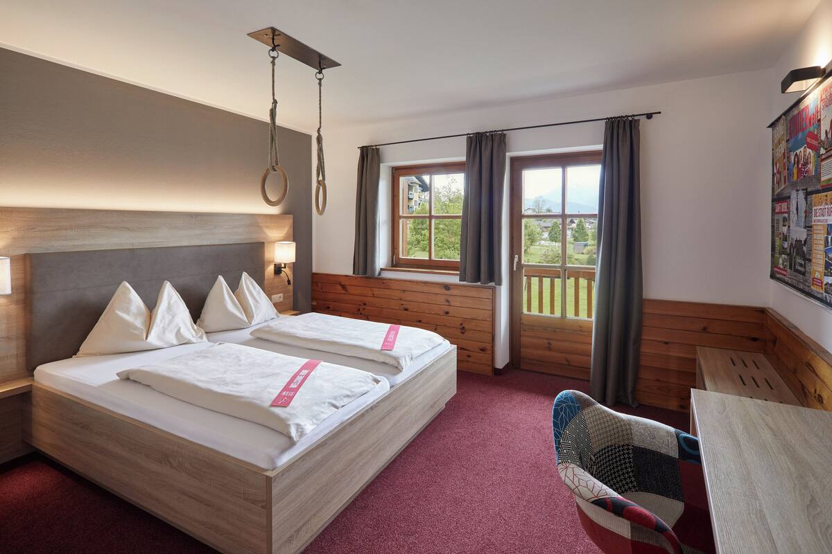 Ein Hotelzimmer mit zwei Betten, einem Holzkopfende, einem Fenster mit Vorhängen, Blick ins Grüne und einer Wandlampe.